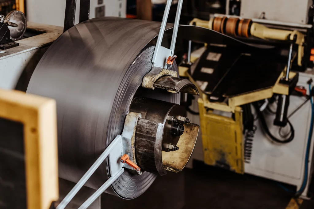 Eine große Metallspule, die auf einer Maschine in einer Industrieumgebung montiert ist, teilweise abgewickelt, mit sichtbaren Rollen und mechanischen Teilen im Hintergrund – ein eindrucksvolles Beispiel für Industriefotografie, das die Präzision und das Ausmaß von Fuchs Metalltechnik einfängt.