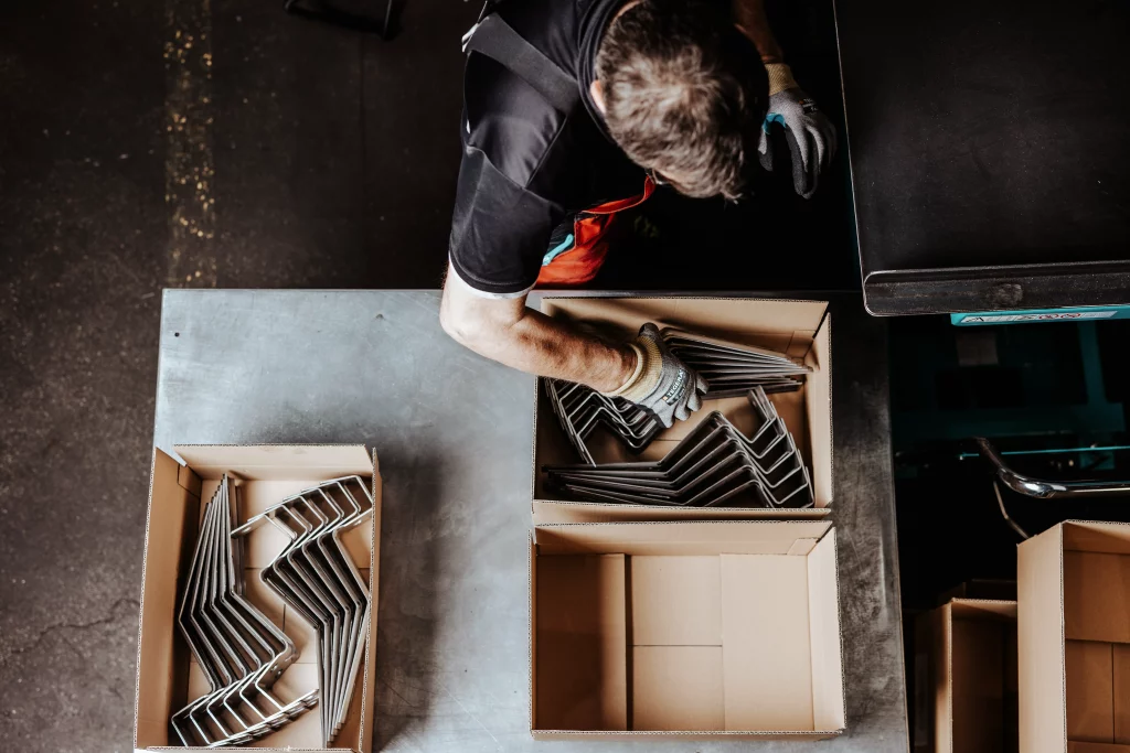 Eine Person, die Handschuhe trägt, sortiert Metallteile auf einem Arbeitstisch in Kartons – aus der Vogelperspektive betrachtet ein eindrucksvolles Beispiel für „Technik trifft Handwerk“, eingefangen durch Industriefotografie.