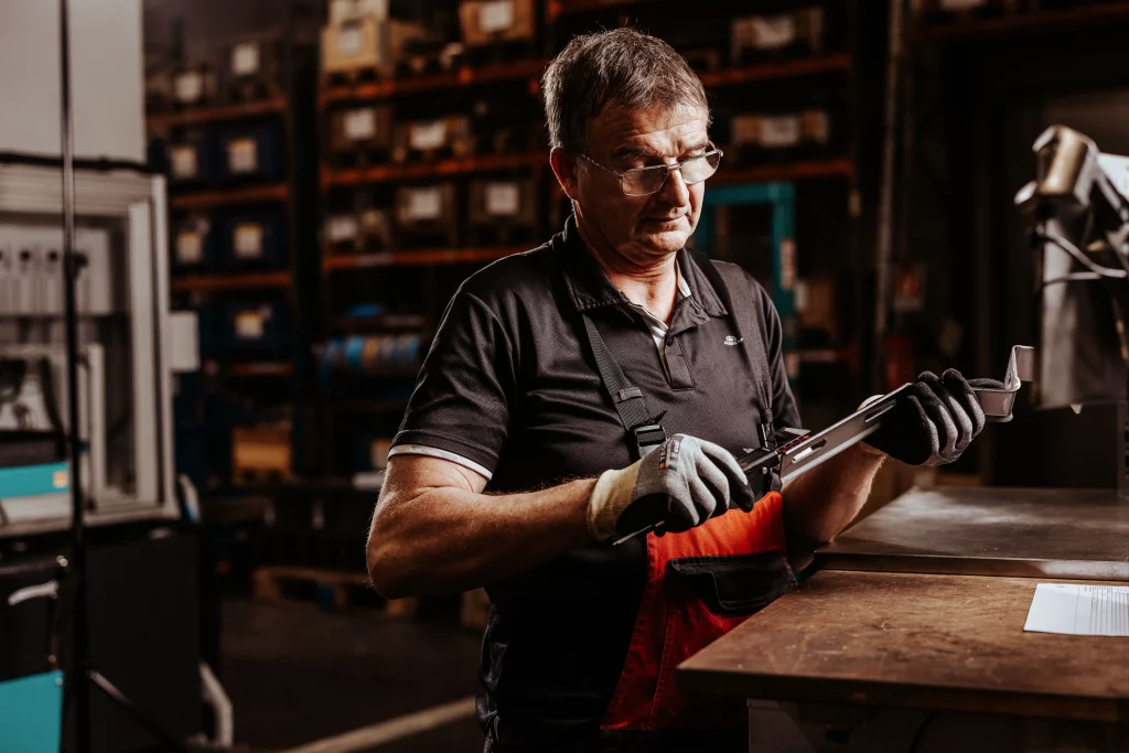 Ein Mann mit Handschuhen und Brille misst mit einem Messschieber ein Metallteil an einer Werkbank in einer Industriewerkstatt, im Hintergrund sind Regale zu sehen – ein authentischer Moment der Industriefotografie, der bei Fuchs Metalltechnik, wo Technik auf Handwerk trifft, perfekt eingefangen wurde.