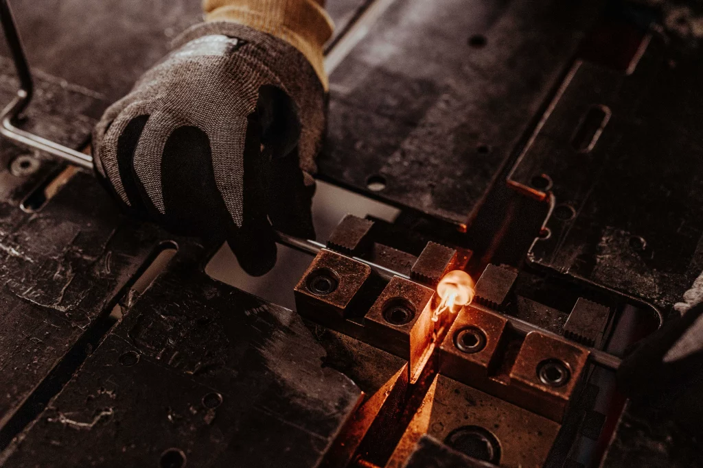 Eine mit einem Handschuh bekleidete Hand hält eine Metallzange über einen Schraubstock, während ein Metallstück an einem Arbeitsplatz bis zur orange leuchtenden Glut erhitzt wird – ein perfektes Beispiel dafür, wie Technik auf Handwerk trifft. Dieser eindrucksvolle Moment, der durch die Industriefotografie von Fuchs Metalltechnik eingefangen wurde, unterstreicht die Präzision und Handwerkskunst, die in der modernen Metallbearbeitung zum Tragen kommen.