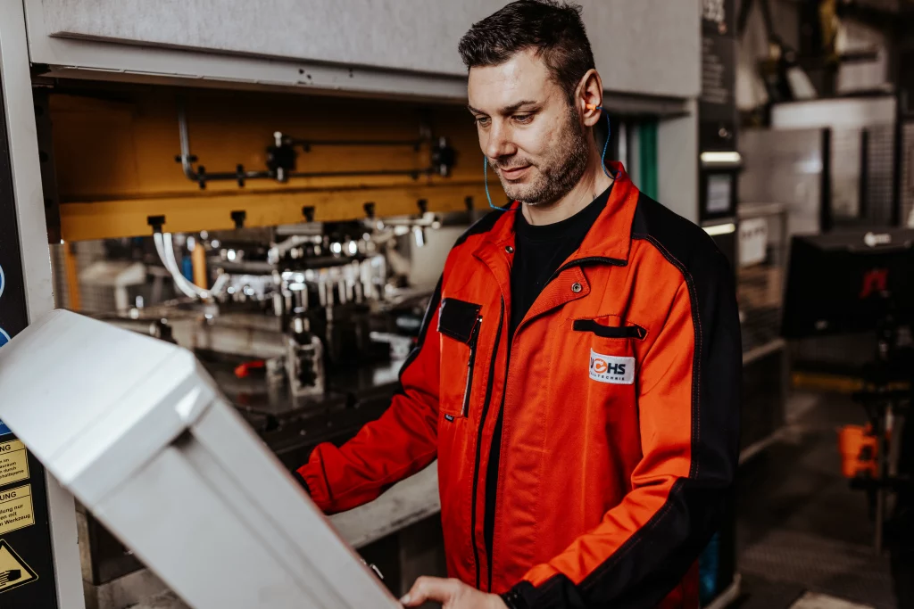 Ein Mann in einer rot-schwarzen Arbeitsjacke bedient ein Bedienpult in einer Industrie- oder Produktionsanlage und verkörpert damit moderne Technik und Präzision, wie sie häufig in der Industriefotografie zu sehen sind.