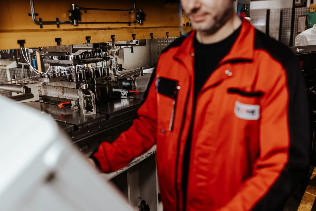 Eine Person in einer roten Arbeitsjacke bedient Maschinen in einer Industrieumgebung bei Fuchs Metalltechnik; auf einer Werkbank sind verschiedene mechanische Teile und Werkzeuge zu sehen, was den Geist der modernen Industriefotografie und innovativer Technik einfängt.