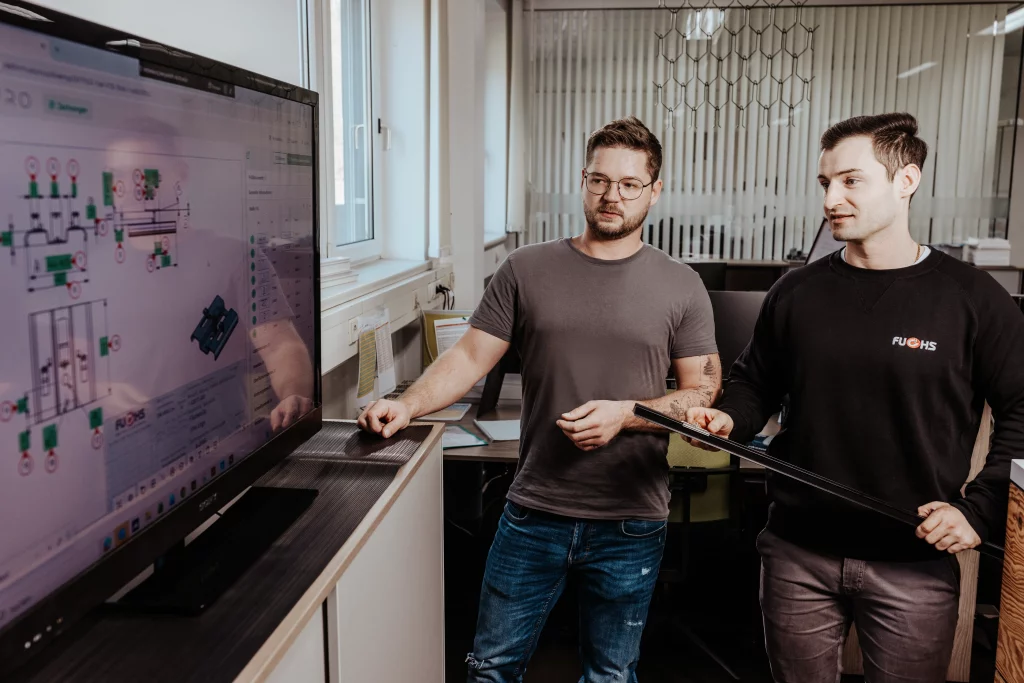 Zwei Männer stehen in einem Büro von Fuchs Metalltechnik und betrachten einen großen Monitor, auf dem technische Diagramme zu sehen sind. Einer der Männer zeigt mit einem Zeigestab auf den Bildschirm und verkörpert damit den Geist der Industriefotografie, in der Technik auf Handwerk trifft.