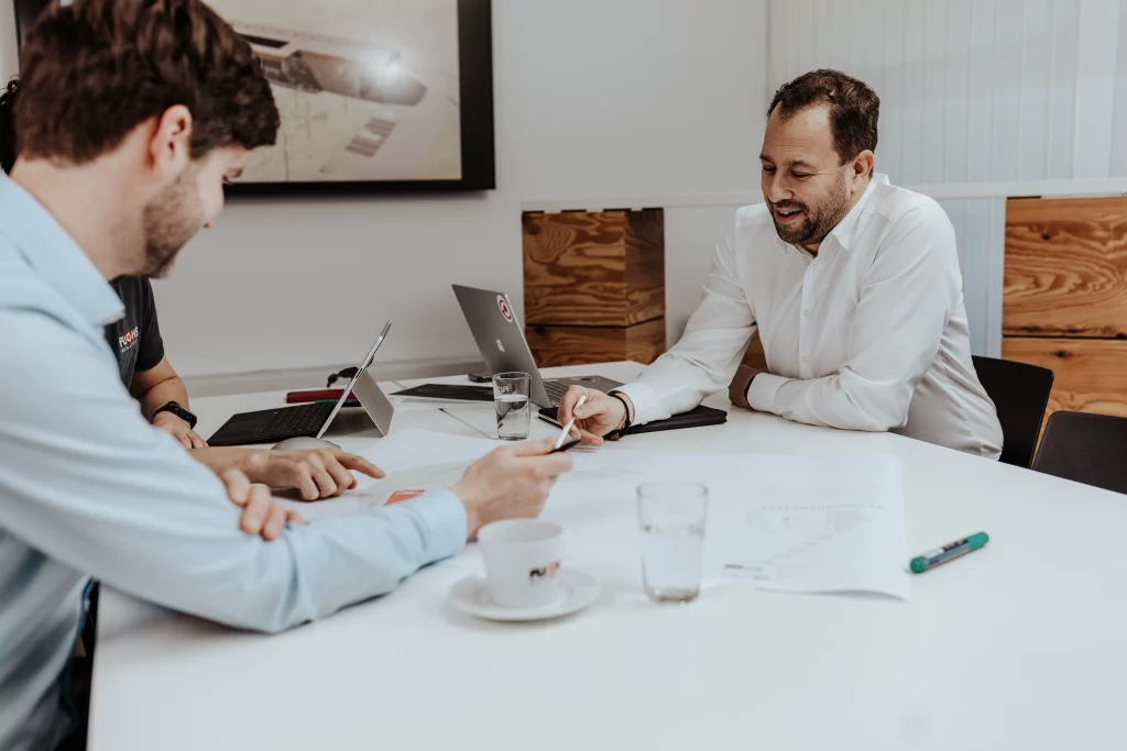 Drei Männer sitzen mit Laptops, Unterlagen und Kaffeetassen um einen weißen Tisch herum und führen in einer modernen Büroumgebung ein Geschäftstreffen – eine perfekte Szene, in der Technik auf Handwerk trifft. Dieses Bild fängt die Essenz der Industriefotografie ein, während Fachleute von Fuchs Metalltechnik gemeinsam an neuen Ideen arbeiten.
