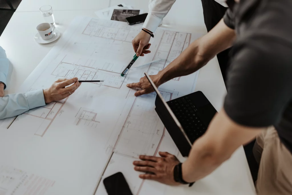 Drei Personen prüfen und zeigen auf Architekturpläne, die auf einem Tisch liegen, auf dem sich ein Laptop, ein Smartphone, eine Tasse und Unterlagen befinden – und fangen so die Essenz der Industriefotografie ein, bei der Technik auf Handwerk trifft. Diese Szene veranschaulicht perfekt den Teamgeist bei Fuchs Metalltechnik und unterstreicht die Schnittstelle zwischen moderner Technologie und handwerklichem Können.