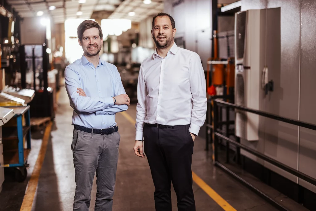 Zwei Männer in Hemden und Anzughosen stehen in einer Industrie- oder Fabrikumgebung, blicken in die Kamera und lächeln, während im Hintergrund Maschinen und Anlagen zu sehen sind – ein perfektes Beispiel für Industriefotografie bei Fuchs Metalltechnik, wo Technik auf Handwerk trifft.