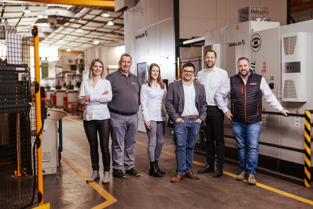 Sechs Personen stehen nebeneinander in einer Fabrik und posieren für ein Gruppenfoto, das den Geist der Industriefotografie einfängt. In Business-Casual-Kleidung verkörpern sie die Synergie von „Technik trifft Handwerk“ bei Fuchs Metalltechnik, während im Hintergrund beeindruckende Industriemaschinen zu sehen sind.