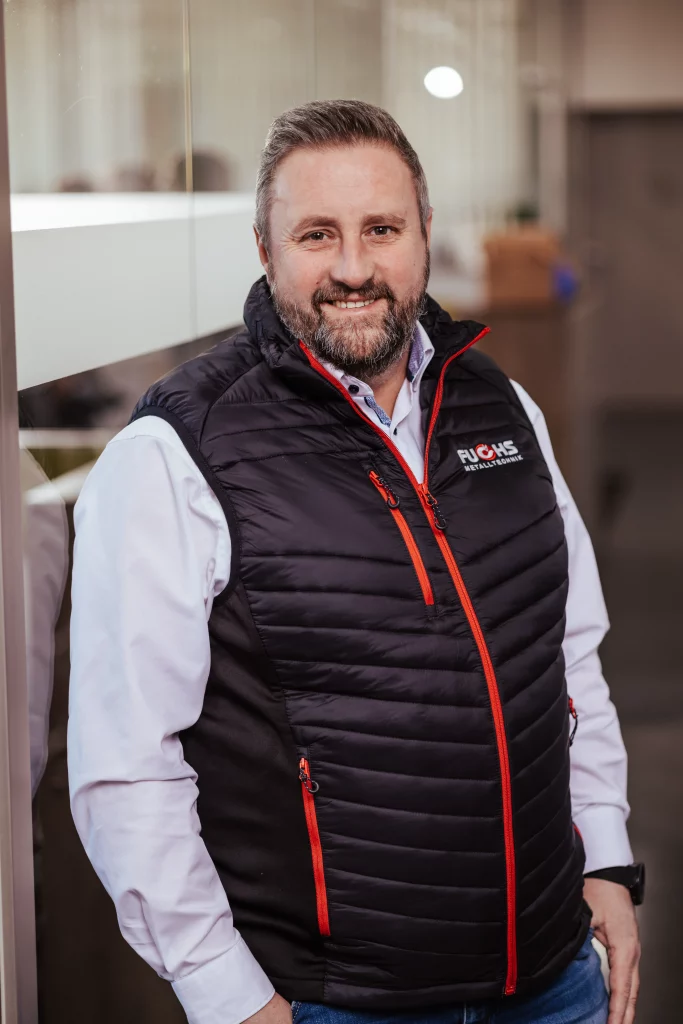 Ein bärtiger Mann in einer schwarzen, gesteppten Weste und einem weißen Hemd steht in einem Innenraum und lächelt in die Kamera – ein perfekter Moment der Industriefotografie, der den Geist von „Technik trifft Handwerk“ bei Fuchs Metalltechnik einfängt.