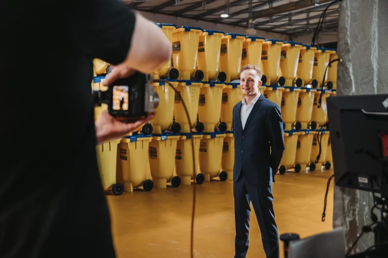 Ein Mann im Anzug posiert für ein Businessfotografie-Foto vor gestapelten gelben Plastikbehältern in einem Lagerhaus am Wiener Ölhafen, während eine andere Person eine Kamera hält.