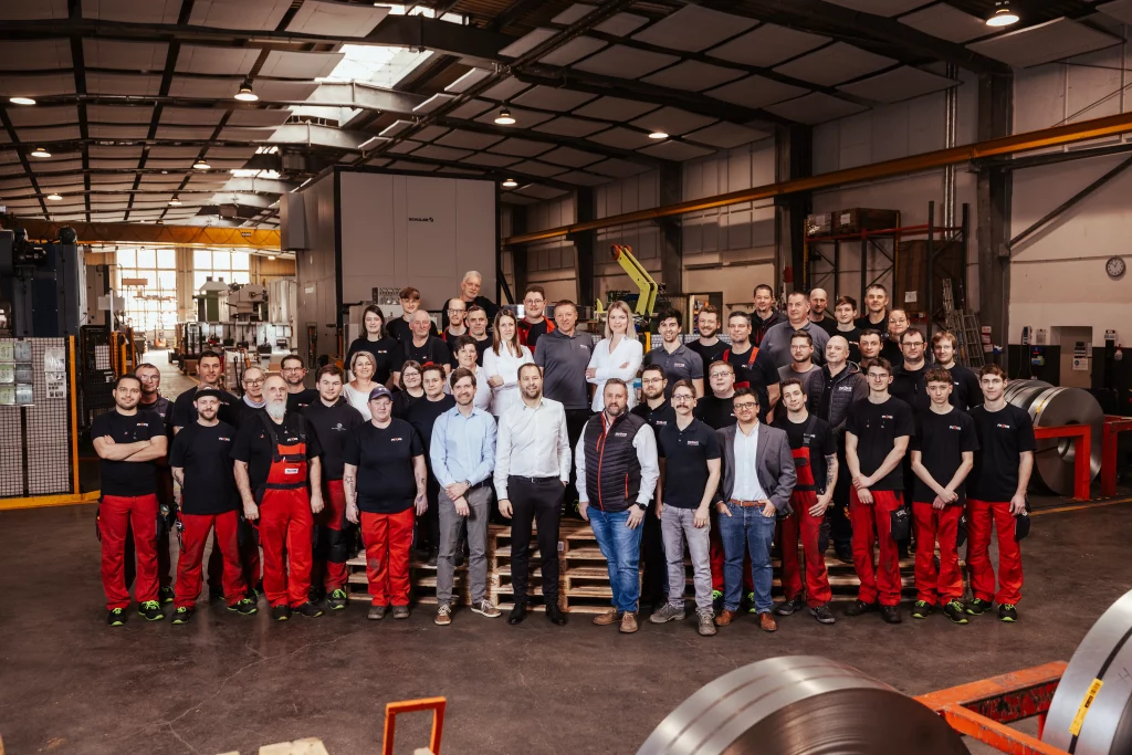 Eine große Gruppe von Menschen in Arbeitskleidung posiert für ein Gruppenfoto in einer weitläufigen Industriehalle, im Hintergrund sind Metallmaschinen zu sehen – ein Bild, das den Geist der Industriefotografie perfekt einfängt. Dieses Bild veranschaulicht auf ideale Weise, wie bei Fuchs Metalltechnik Technik auf Handwerk trifft.