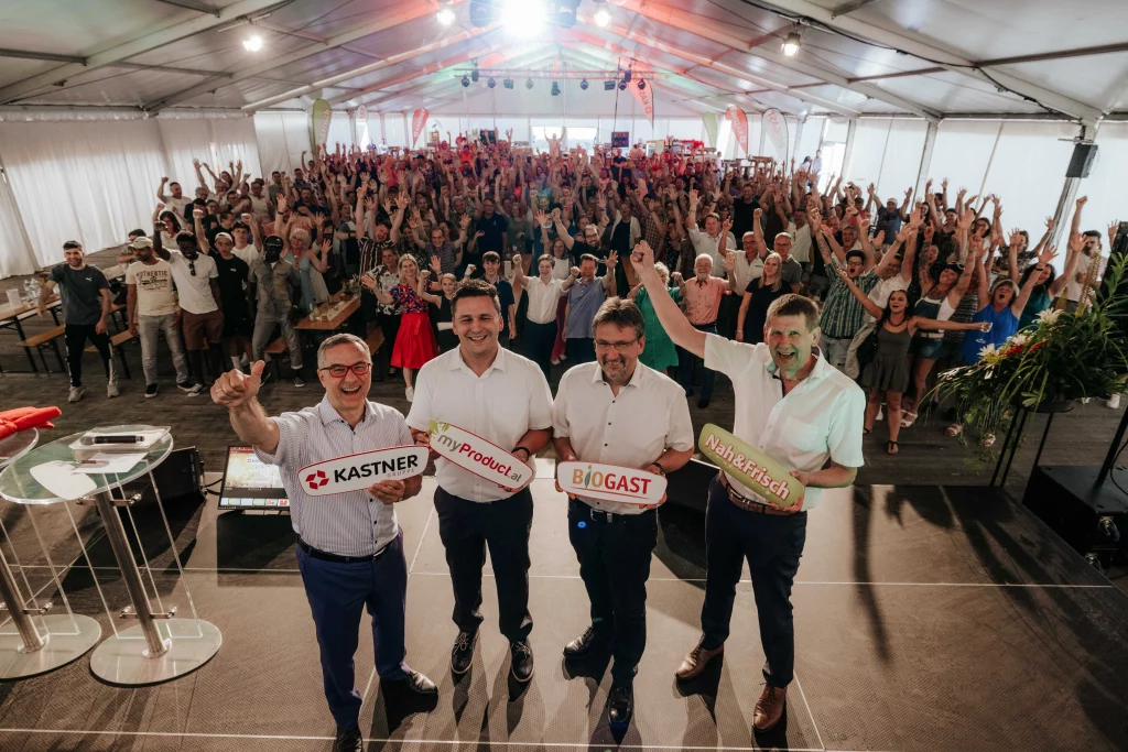 Vier Männer auf der Bühne halten Schilder mit Firmenlogos vor einem großen, begeisterten Publikum in einem Veranstaltungszelt hoch und halten dabei „echte Momente“ lebendiger Eventfotografie für die KASTNER Gruppe fest.