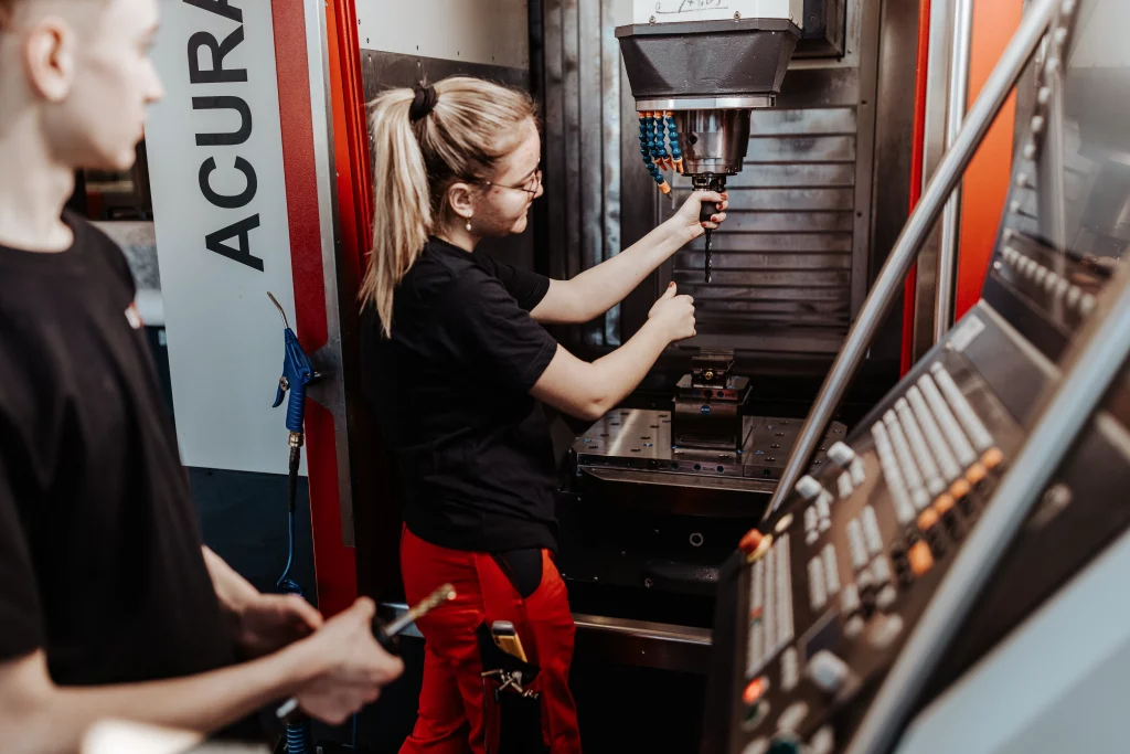 Zwei Personen bedienen eine CNC-Maschine; eine justiert den Werkzeugkopf, während die andere die Ausrüstung festhält. Im Hintergrund sind Bedienfelder und Maschinen in einer industriellen Werkstatteinrichtung zu sehen. Diese Szene veranschaulicht das Motto „Technik trifft Handwerk“ und fängt den Geist der Industriefotografie bei Fuchs Metalltechnik ein.