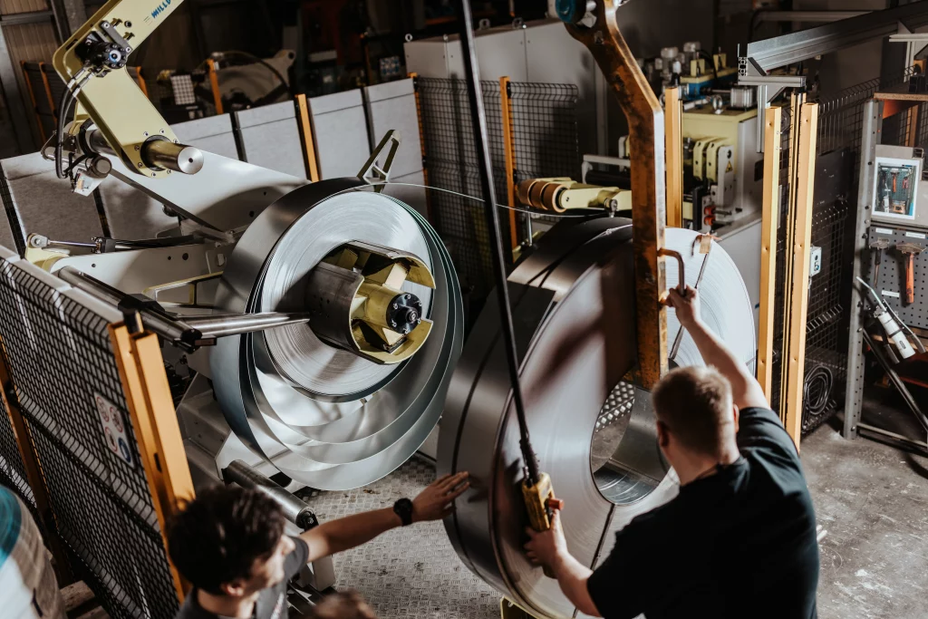 Zwei Mitarbeiter bedienen Maschinen zur Handhabung großer Blechrollen in einer industriellen Fertigungsumgebung, in der „Technik trifft Handwerk“ und „Fuchs Metalltechnik“ für höchste Präzision sorgen.