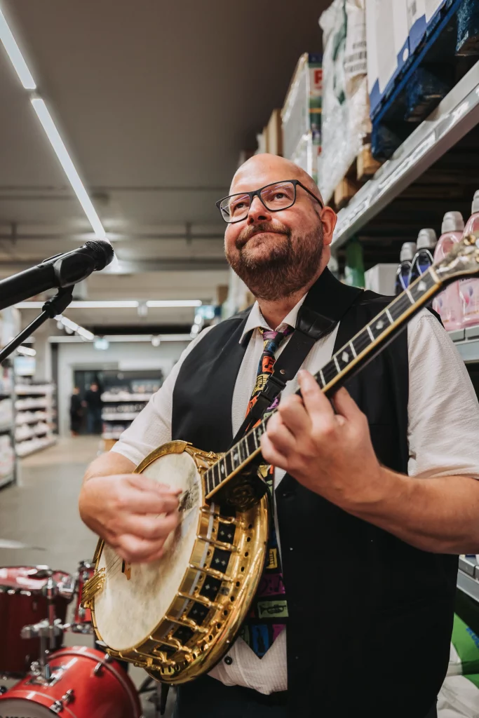 Ein Mann mit Brille spielt Banjo und singt in ein Mikrofon, während im Hintergrund ein Laden gang mit Regalen und Produkten der KASTNER Gruppe zu sehen ist – so entstehen echte Momente, die sich perfekt für die Eventfotografie eignen.