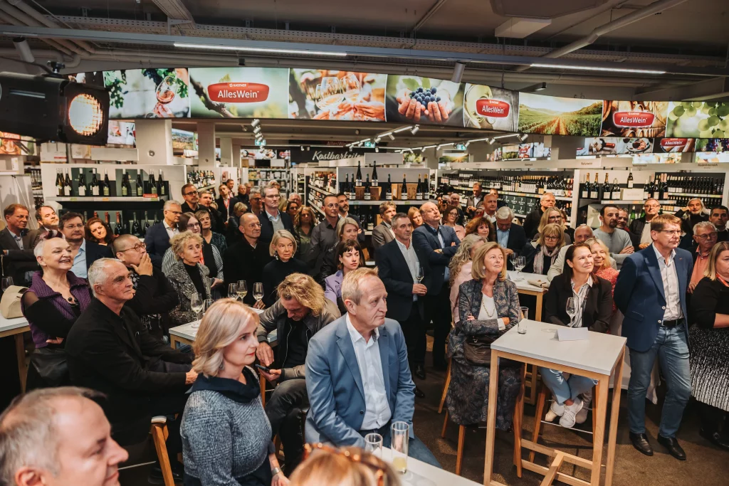 Eine große Gruppe von Erwachsenen lauscht aufmerksam während einer Veranstaltung in einem Geschäft, im Hintergrund sind Regale mit Wein und Lebensmitteln zu sehen. Dabei werden „echte Momente“ durch professionelle Eventfotografie festgehalten – organisiert von der KASTNER Gruppe.