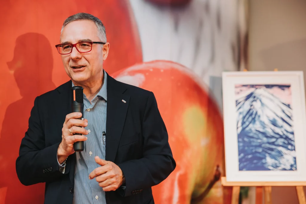 Ein Mann im Anzug und mit Brille spricht in ein Mikrofon, während er in einem Raum neben einem gerahmten Bild eines Berges auf einer Staffelei steht – eine ideale Szene „echter Momente“, eingefangen durch Eventfotografie bei einer Veranstaltung der KASTNER Gruppe.