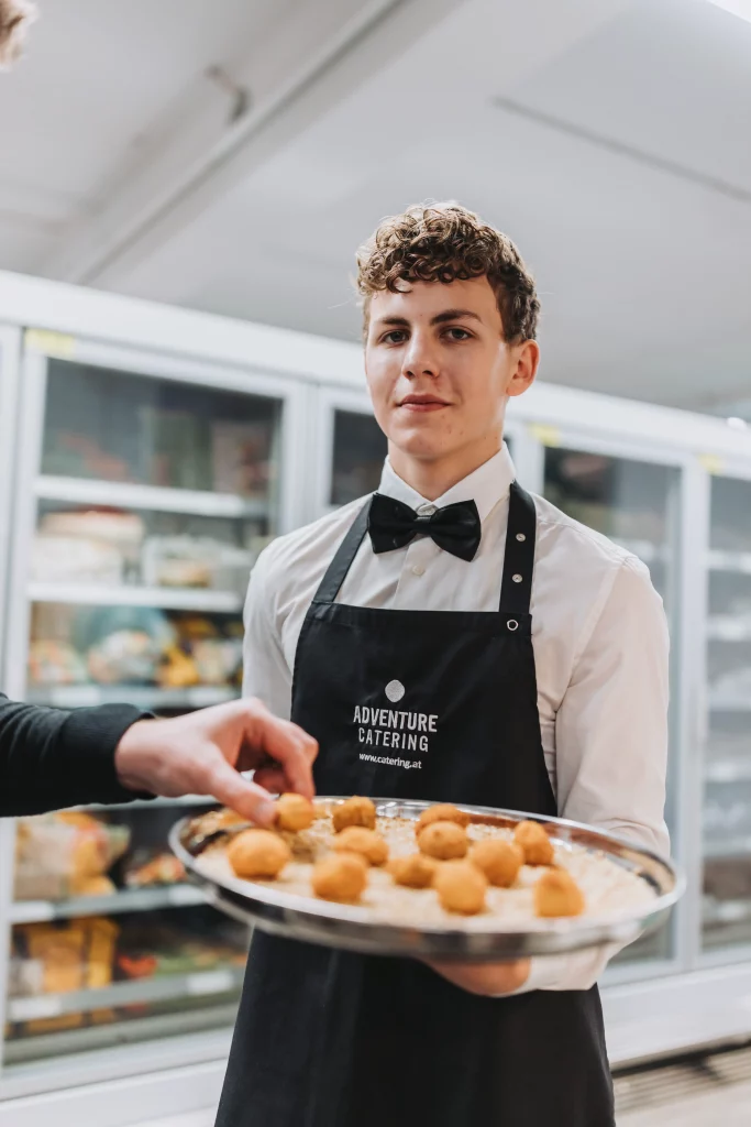 Ein Kellner mit Fliege und einer Schürze von „Adventure Catering“ hält ein Tablett mit Häppchen in der Hand, während jemand eines davon nimmt – so entstehen „echte Momente“ vor einer Kulisse mit Kühlschränken im Hintergrund – perfekt für authentische Eventfotografie der KASTNER Gruppe.