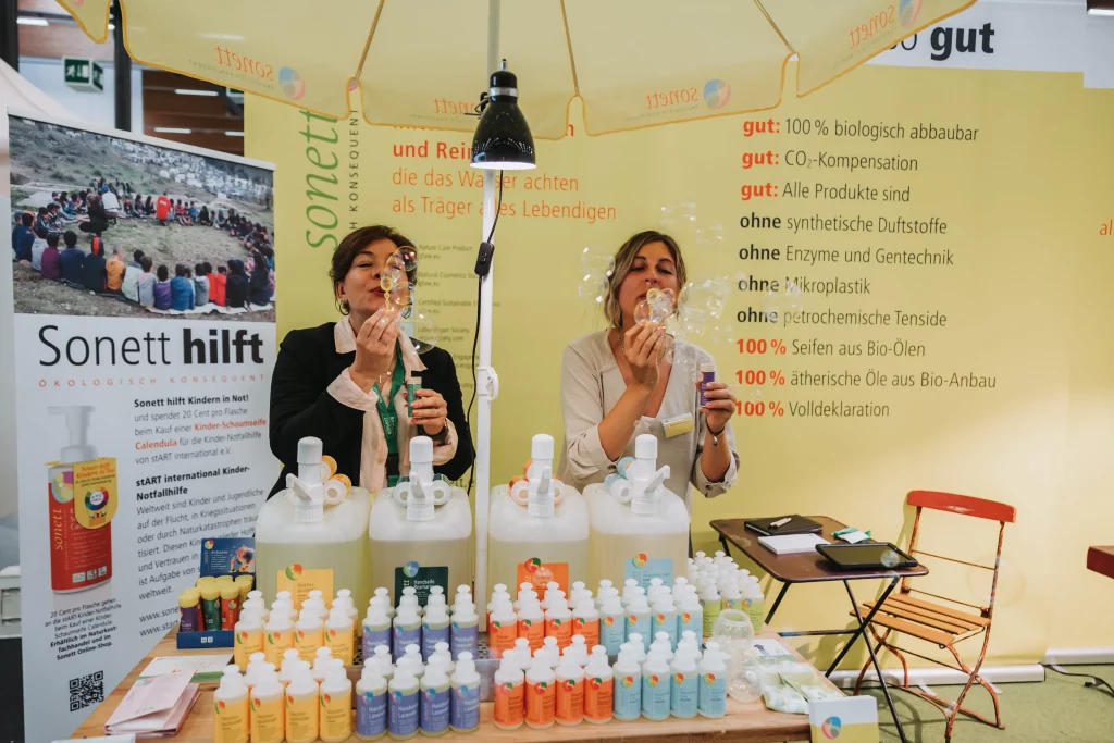 Zwei Frauen sitzen an einem Stand mit Reinigungsmitteln, lassen Seifenblasen steigen und genießen „echte Momente“. Hinter ihnen sind ein gelber Regenschirm und Werbebanner zu sehen. Auf dem Tisch stehen Flaschen in verschiedenen Größen, festgehalten in lebendiger Eventfotografie für die KASTNER Gruppe.