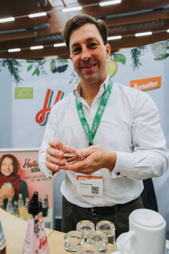 Ein Mann in einem weißen Hemd hält ein Glas in der Hand und lächelt an einem Stand, an dem Schilder der KASTNER Gruppe zu sehen sind und vor ihm auf dem Tisch Produkte ausgestellt sind – so werden durch Eventfotografie echte Momente festgehalten.