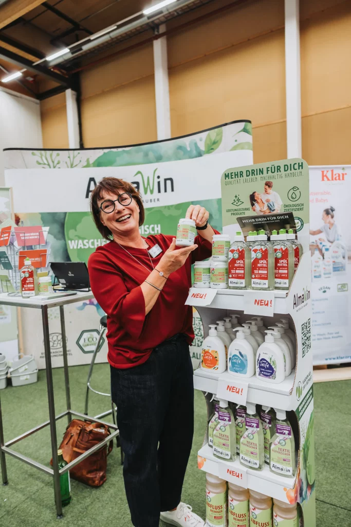 Eine Frau in einem roten Oberteil steht in einem Raum, lächelt und hält eine Flasche Waschmittel neben einem Regal mit AlmaWin-Reinigungsprodukten in der Hand – eine authentische Momentaufnahme, die „echte Momente“ perfekt einfängt, wie sie für die Eventfotografie der KASTNER Gruppe typisch sind.