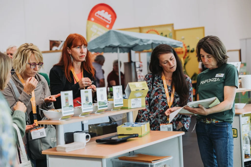 Vier Frauen stehen an einem Ausstellungsstand mit Produkten, lesen eine Broschüre und unterhalten sich; im Hintergrund sind weitere Personen und Stände zu sehen. Diese Schnappschussaufnahme fängt echte Momente einer Veranstaltung der KASTNER-Gruppe ein und zeigt authentische Interaktionen, wie sie für hochwertige Eventfotografie typisch sind.