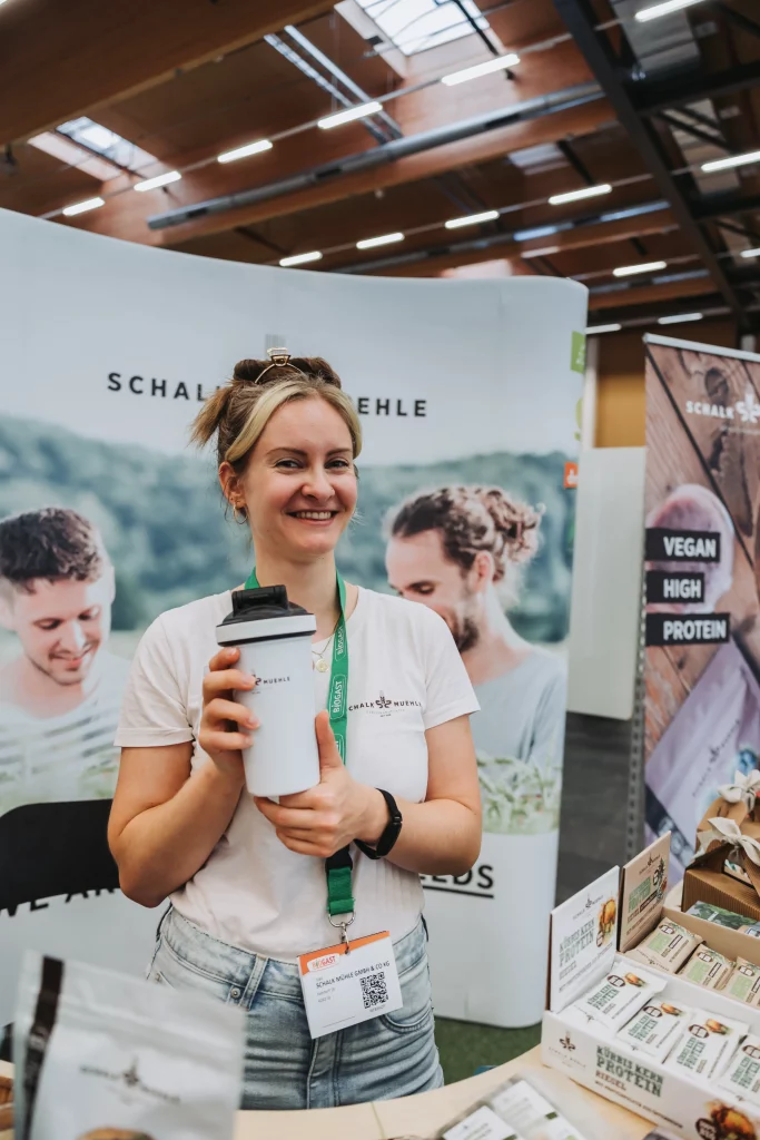Eine Frau steht an einem Stand, hält einen Mehrwegbecher in der Hand, trägt ein Namensschild und lächelt, hinter ihr sind Werbebanner und Produktpräsentationen der KASTNER Gruppe zu sehen – so entstehen „echte Momente“ der Eventfotografie.