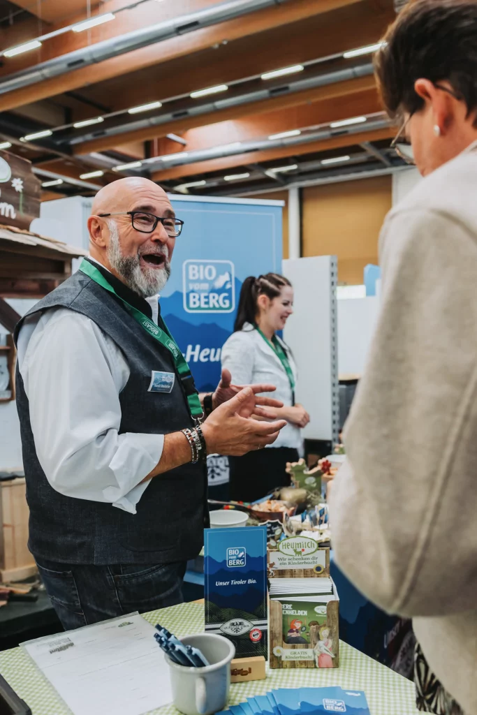 Ein älterer Mann mit Brille unterhält sich mit einem Besucher am Messestand der KASTNER Gruppe, während eine Frau daneben steht. Auf dem Tisch sind Werbematerialien und Produkte ausgestellt, die durch professionelle Eventfotografie echte Momente der Veranstaltung einfangen.