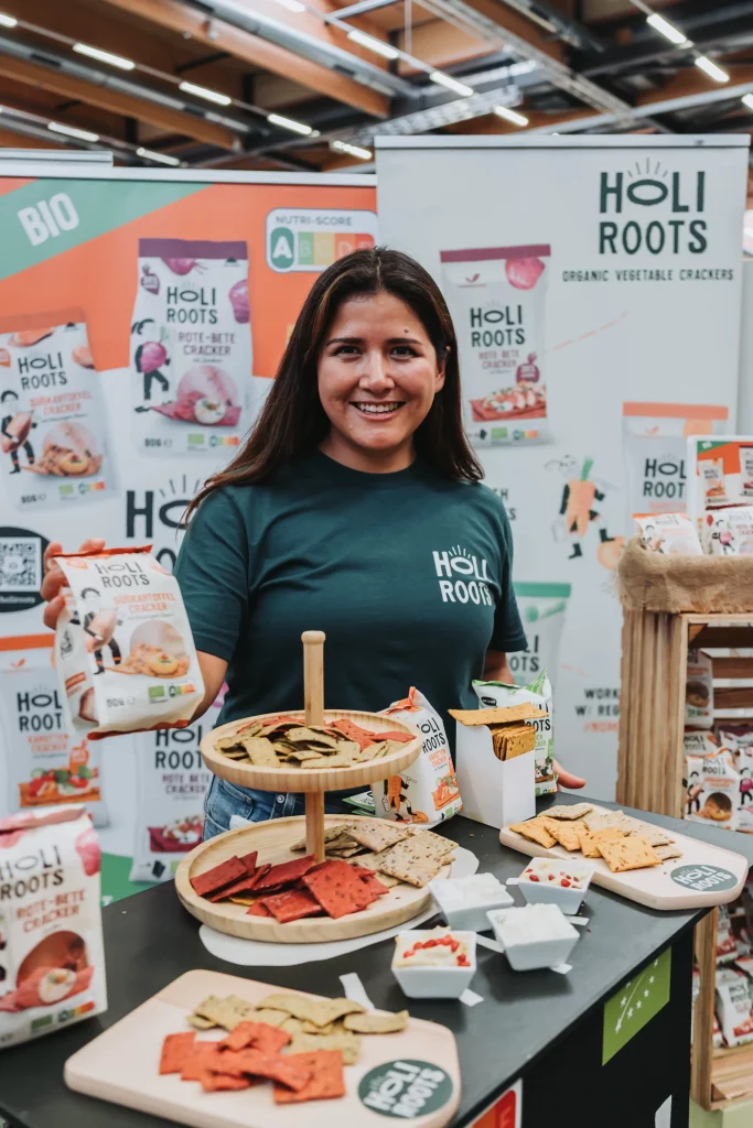 Eine Frau steht an einem Stand, an dem Bio-Gemüsecracker von Holi Roots ausgestellt sind. Sie hält eine Produktverpackung in der Hand und lächelt, während auf dem Tisch Kostproben und Produktverpackungen liegen – so entstehen „echte Momente“ lebendiger Eventfotografie für die KASTNER Gruppe.
