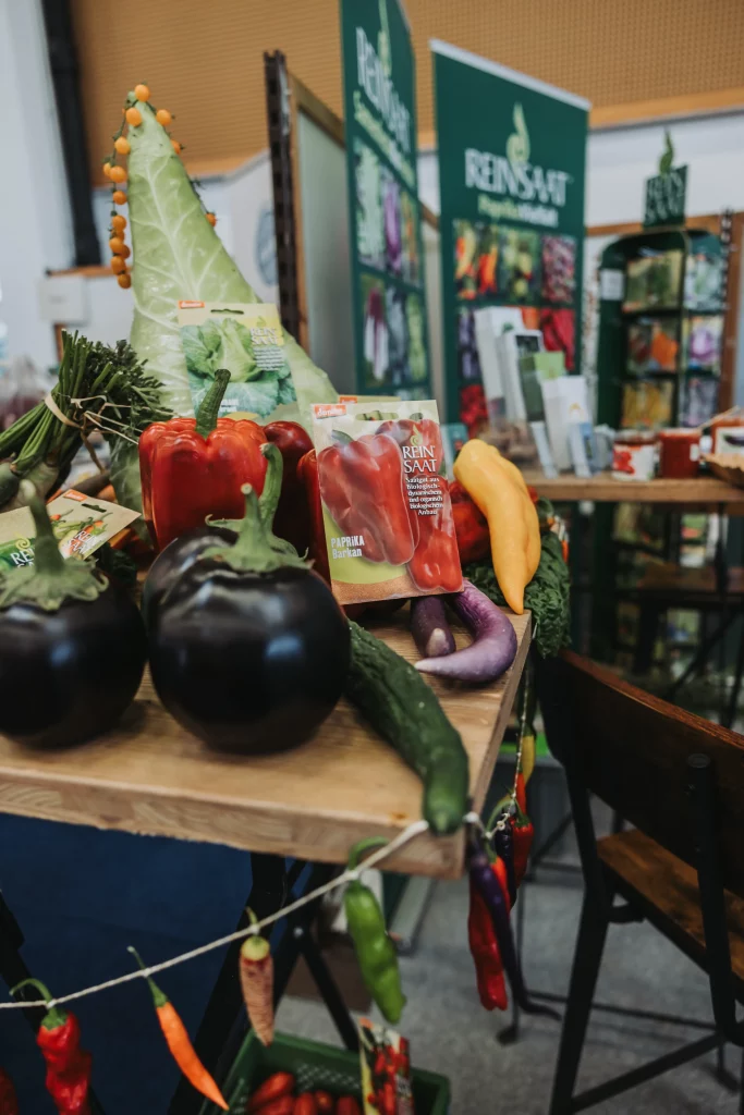 Ein Ausstellungsstand mit frischem Gemüse wie Auberginen, Paprika und Gurken sowie Saatgutpäckchen auf einem Hallenmarkt oder an einem Messestand – festgehalten durch professionelle Eventfotografie der KASTNER Gruppe, um authentische Momente mit regionalen Erzeugnissen in den Vordergrund zu rücken.