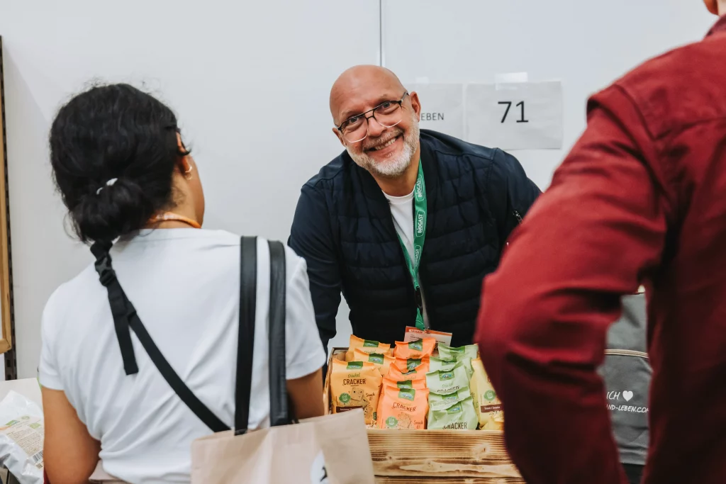 Eine Person lächelt hinter einem Tisch, auf dem Snackpackungen ausgestellt sind, und unterhält sich mit Besuchern an einem Stand mit der Nummer 71 – als Teil der ansprechenden Eventfotografie der KASTNER Gruppe.