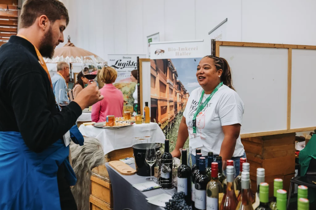 Eine Frau steht hinter einem Stand mit Wein und Honig und unterhält sich mit einem Mann, der ein Getränk in der Hand hält, auf einem Hallenmarkt oder einer Messe. Solche authentischen Momente einzufangen, gehört zur Eventfotografie der KASTNER-Gruppe.