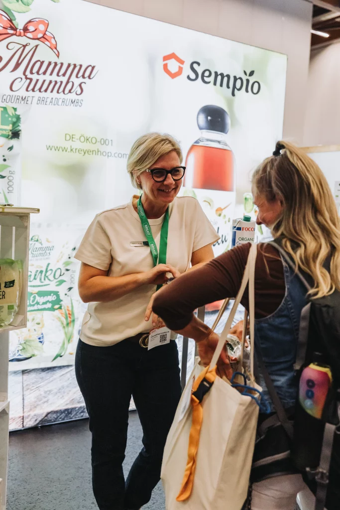 Zwei Frauen unterhalten sich an einem Messestand, im Hintergrund sind die Marken „Mamma Crumbs“ und „Sempio“ zu sehen. Diese „echten Momente“, eingefangen durch Eventfotografie: Eine der Frauen trägt ein Namensschild und lächelt, während sie mit der anderen spricht – ein perfektes Beispiel für den Geist des Netzwerks bei einer Veranstaltung der KASTNER Gruppe.