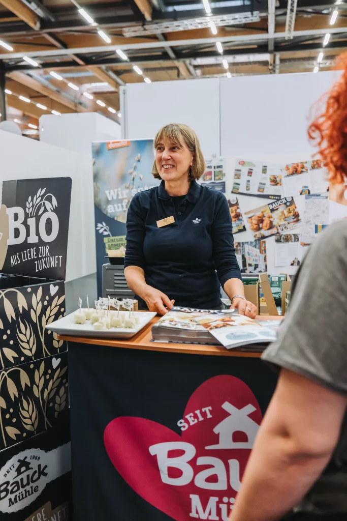 Eine Frau steht hinter einem Messestand und unterhält sich lächelnd mit den Besuchern. Am Stand wird Werbematerial für Bio-Produkte der KASTNER Gruppe präsentiert, das durch professionelle Eventfotografie perfekt in Szene gesetzt wird.