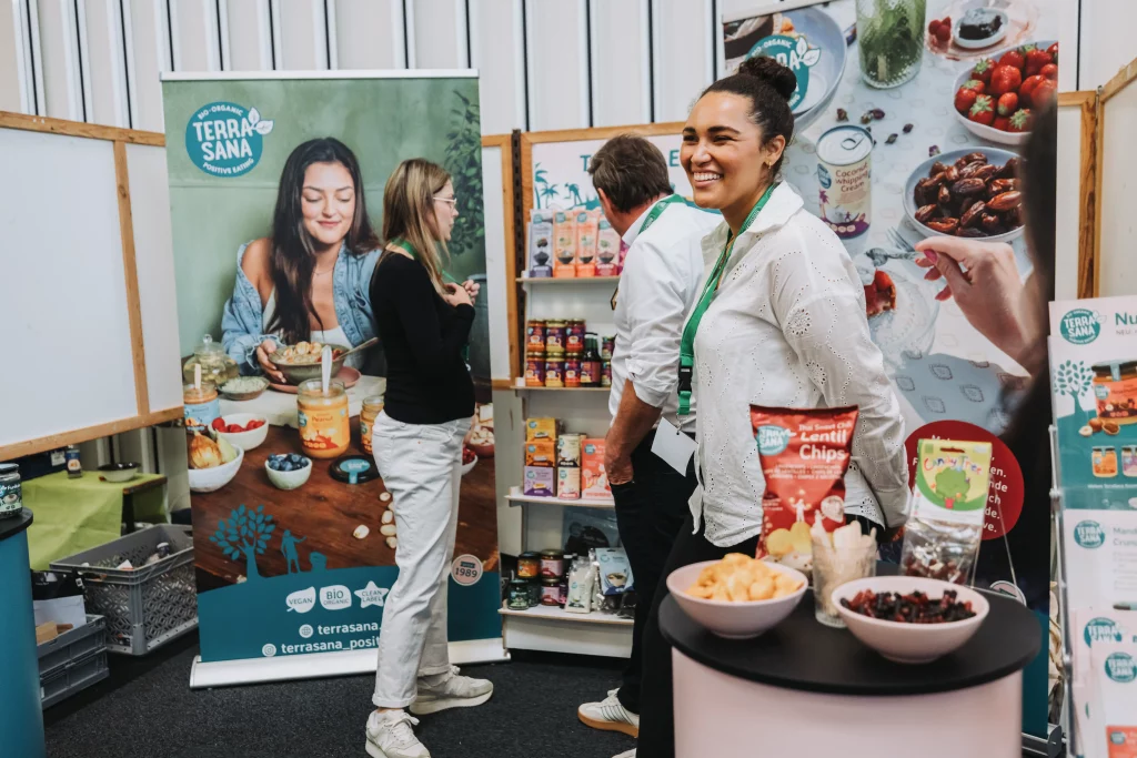Drei Personen unterhalten sich an einem Messestand, an dem Snacks und Werbematerialien der Marke TerraSana präsentiert werden; die Produkte sind übersichtlich in Regalen und auf Tischen angeordnet. Diese Szene aus der Eventfotografie fängt „echte Momente“ ein und unterstreicht die natürliche Interaktion sowie die dynamische Atmosphäre des Auftritts der KASTNER Gruppe.