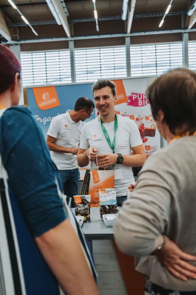 Ein Mann in einem weißen Hemd unterhält sich mit Besuchern an einem Messestand, auf dessen Theke Werbematerialien und Snacks ausliegen, und hält dabei echte Momente des Netzwerks und der Interaktion fest – ein Beispiel für authentische Eventfotografie für die KASTNER Gruppe.