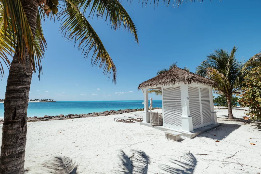 Eine kleine weiße Strandhütte mit Strohdach am Sandstrand, umgeben von Palmen, mit Blick auf das ruhige türkisfarbene Meer unter einem strahlend blauen Himmel – eine idyllische Kulisse, perfekt für Imageproduktion.