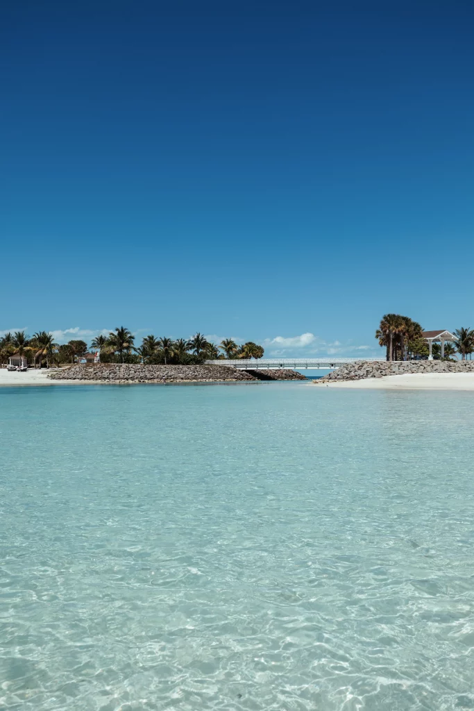 Kristallklares türkisfarbenes Wasser im Vordergrund, ein von Palmen gesäumter Sandstrand und ein kleines Gebäude unter strahlend blauem Himmel zeigen die beeindruckenden Ergebnisse der professionellen Bildproduktion.