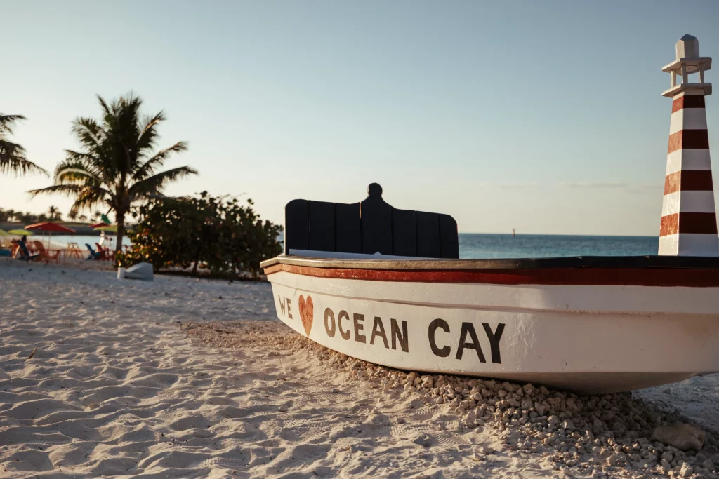 Ein weißes Boot mit der Aufschrift „We ♥ Ocean Cay“ liegt an einem Sandstrand in der Nähe eines Leuchtturms – aufgenommen von Imageproduktion, mit Palmen und dem Meer im Hintergrund.