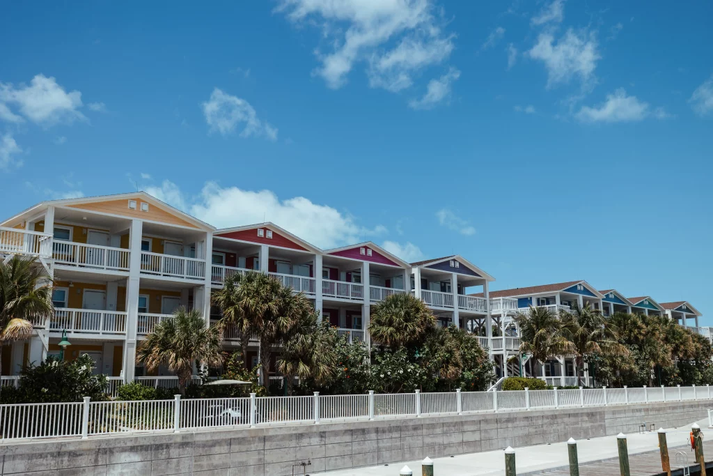 Eine Reihe farbenfroher, zweistöckiger Strandhäuser, die mit professionellen Bildproduktionstechniken erstellt wurden; jedes davon verfügt über einen Balkon, Palmen und einen weißen Zaun vor einem strahlend blauen Himmel mit vereinzelten Wolken.