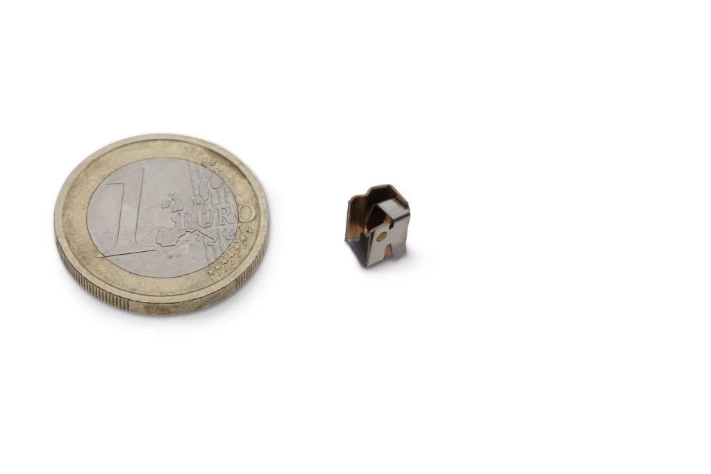 Eine 1-Euro-Münze neben einem kleinen, metallischen, geometrischen Objekt auf weißem Hintergrund, wodurch der Größenvergleich deutlich wird – Industriefotografie vom Feinsten, wo Technik auf Handwerk trifft, von Fuchs Metalltechnik.