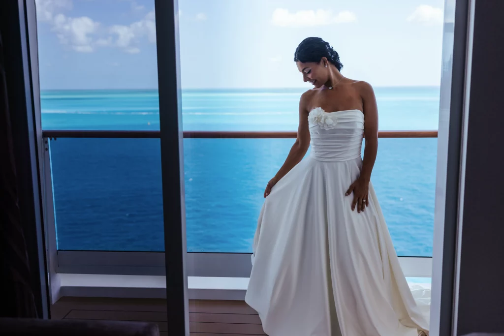 Eine Frau in einem weißen, trägerlosen Brautkleid steht auf einem Balkon mit Blick auf das ruhige blaue Meer und den klaren Himmel – eine elegante Kulisse, die sich perfekt für die Bildproduktion eignet.