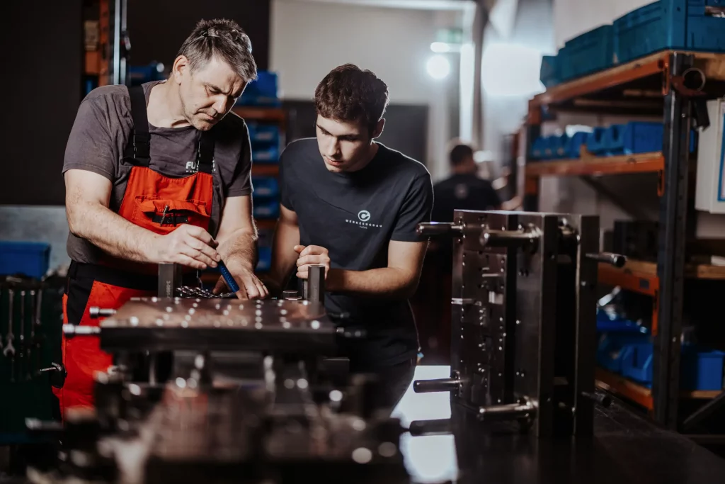 Zwei Männer arbeiten gemeinsam an einer Metallbaugruppe in einer Werkstatt, umgeben von Werkzeugen und Regalen mit blauen Aufbewahrungsbehältern – eine Szene, in der Technik auf Handwerk trifft, perfekt eingefangen in authentischer Industriefotografie bei Fuchs Metalltechnik.