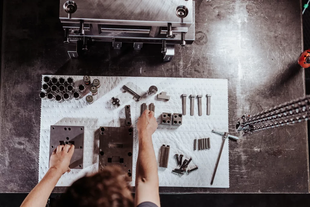 Eine Person, die auf einer gepolsterten Arbeitsfläche verschiedene Metallteile und Werkzeuge für Maschinen zusammenbaut oder prüft, von oben betrachtet – ein präziser Moment der Technik, eingefangen im Stil echter Industriefotografie.