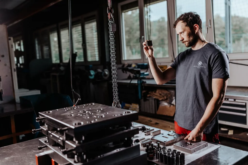 Ein Mann in einer Werkstatt begutachtet ein kleines Metallteil, während er neben großen Metallformen und Werkzeugen an einer Werkbank steht – die Industriefotografie bei Fuchs Metalltechnik dokumentiert eindrucksvoll, wie Technik auf Handwerk trifft.