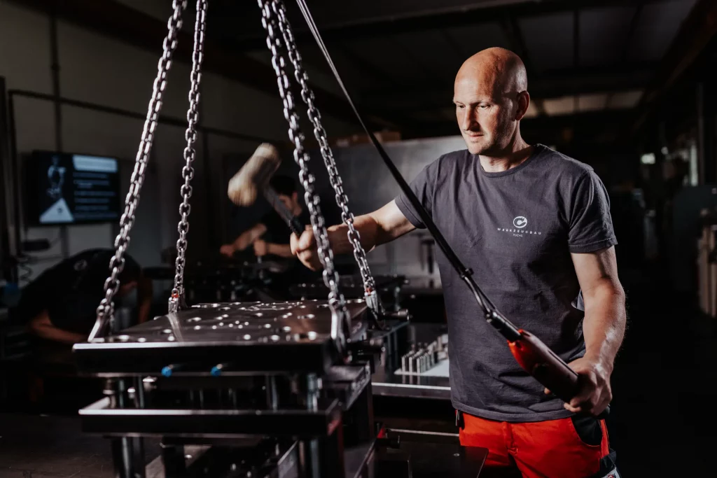Ein Mann bedient Maschinen in einer Werkstatt und bearbeitet mit einem Hammer ein an Ketten aufgehängtes Metallstück – eine Szene, die die Präzision und das Fachwissen, die für die Technik- und Industriefotografie bei Fuchs Metalltechnik von zentraler Bedeutung sind, perfekt zum Ausdruck bringt.