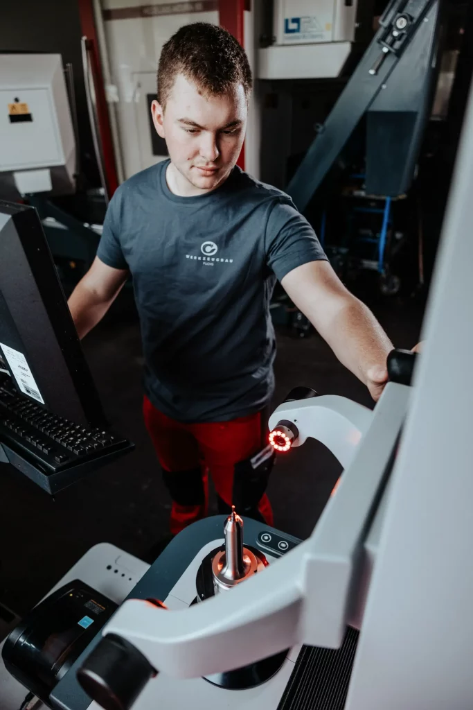 Eine Person bedient eine CNC-Maschine, passt die Einstellungen auf einem Touchscreen an und beobachtet dabei ein Werkzeug, das von einem automatisierten Roboterarm in einer industriellen Umgebung gehalten wird – und fängt so die Präzision und Innovation ein, die für die führende Technik von Fuchs Metalltechnik in der zeitgenössischen Industriefotografie charakteristisch sind.
