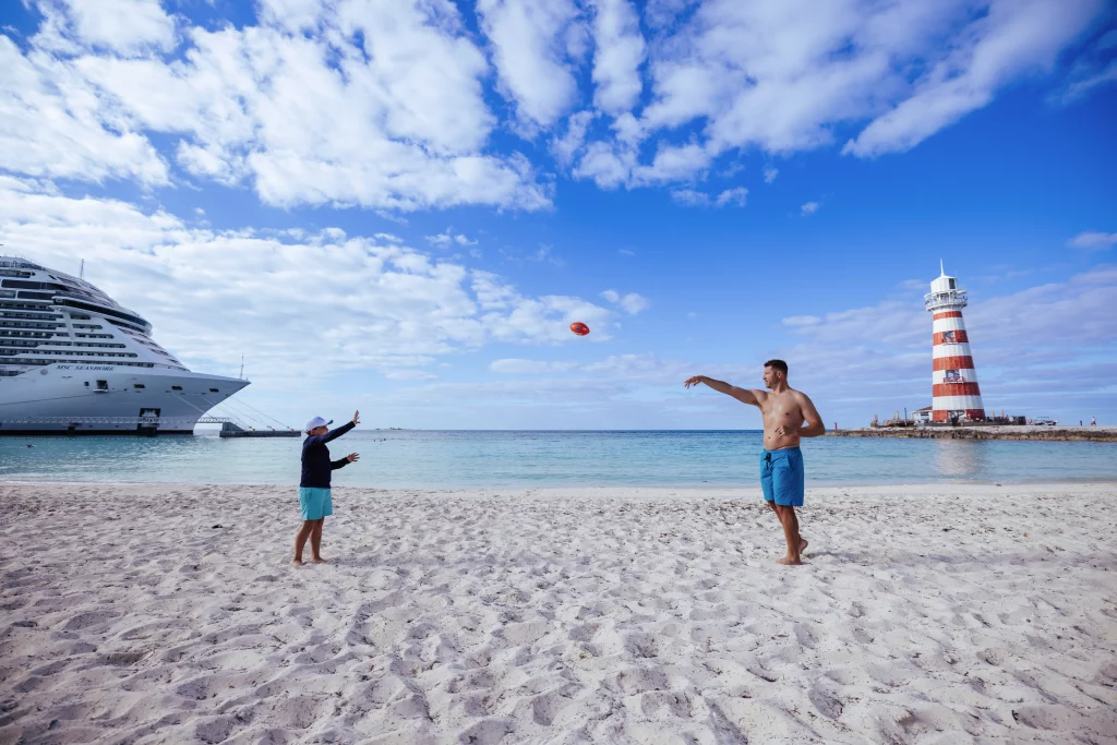 Zwei Menschen spielen an einem Sandstrand mit einem Frisbee, eingefangen in dieser atemberaubenden Bildproduktion. Im Hintergrund sind unter einem blauen Himmel mit Wolken ein Kreuzfahrtschiff und ein rot-weiß gestreifter Leuchtturm zu sehen.