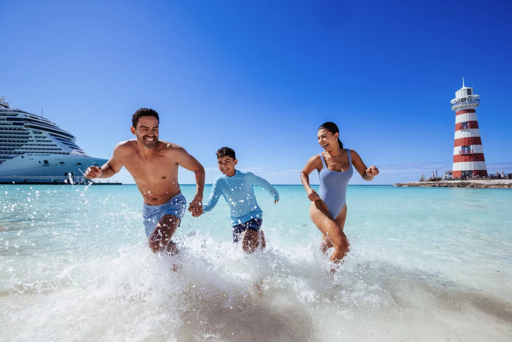 Ein Mann, eine Frau und ein Kind laufen unter strahlend blauem Himmel durch seichtes Wasser an einem Strand in der Nähe eines großen Kreuzfahrtschiffes und eines rot-weiß gestreiften Leuchtturms und halten dabei den perfekten Moment für eine Bildproduktion fest.