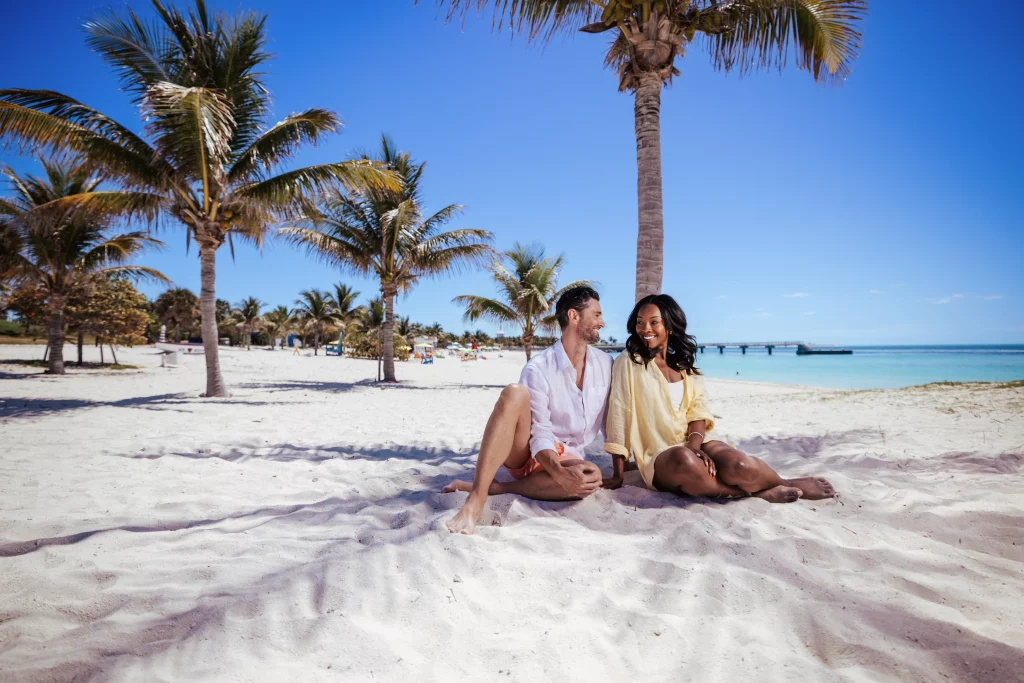 Ein Mann und eine Frau sitzen gemeinsam an einem Sandstrand unter Palmen, lächeln und schauen sich an, im Hintergrund das Meer und der blaue Himmel – eine perfekte Szene für eine Bildproduktion, die die Essenz von Entspannung und Verbundenheit einfängt.