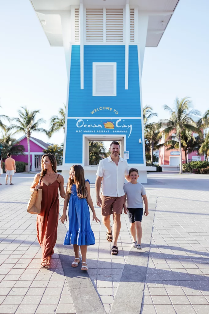 Eine vierköpfige Familie spaziert vor einem blauen „Welcome to Ocean Cay“-Schild vorbei, im Hintergrund sind Palmen und bunte Gebäude zu sehen – ein wunderschönes Bild, das von Imageproduktion meisterhaft eingefangen wurde.